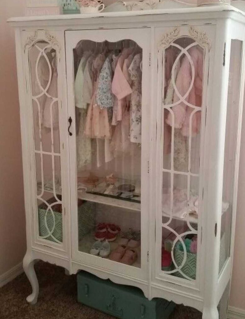 picture of a repurposed vintage white wardrobe with glass in front. inside the wardrobe are several outfits hung up to store a baby's closet items