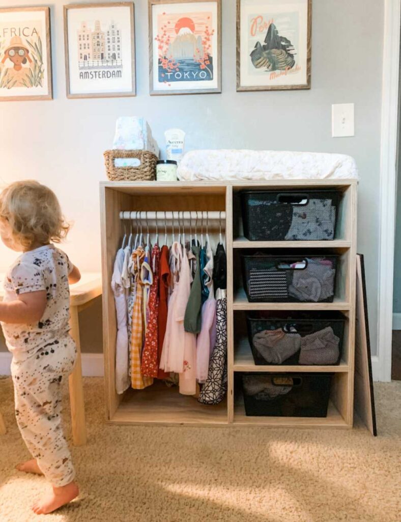 picture of a montessori type open shelving wardrobe and clothing rod with a changing table and diaper supplies on top. To the left is a young child playing 