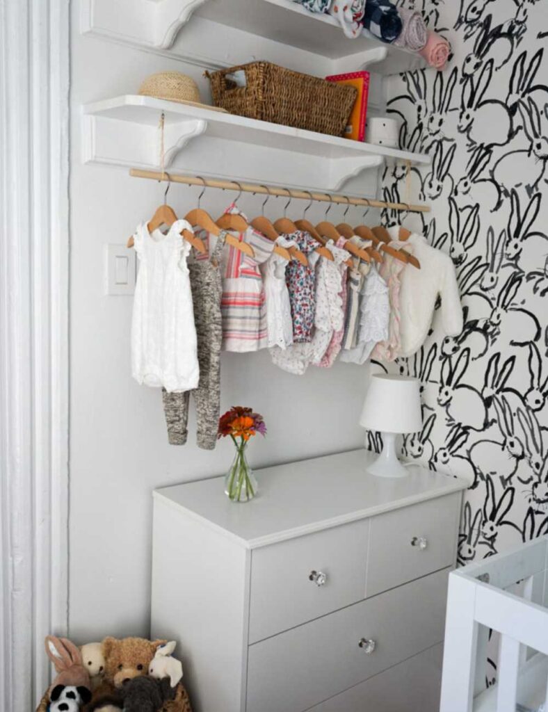 picture showing corner of baby's room with a white dresser and a shelf above it with a clothing rod attached. Several clothing items are hanging on rod.