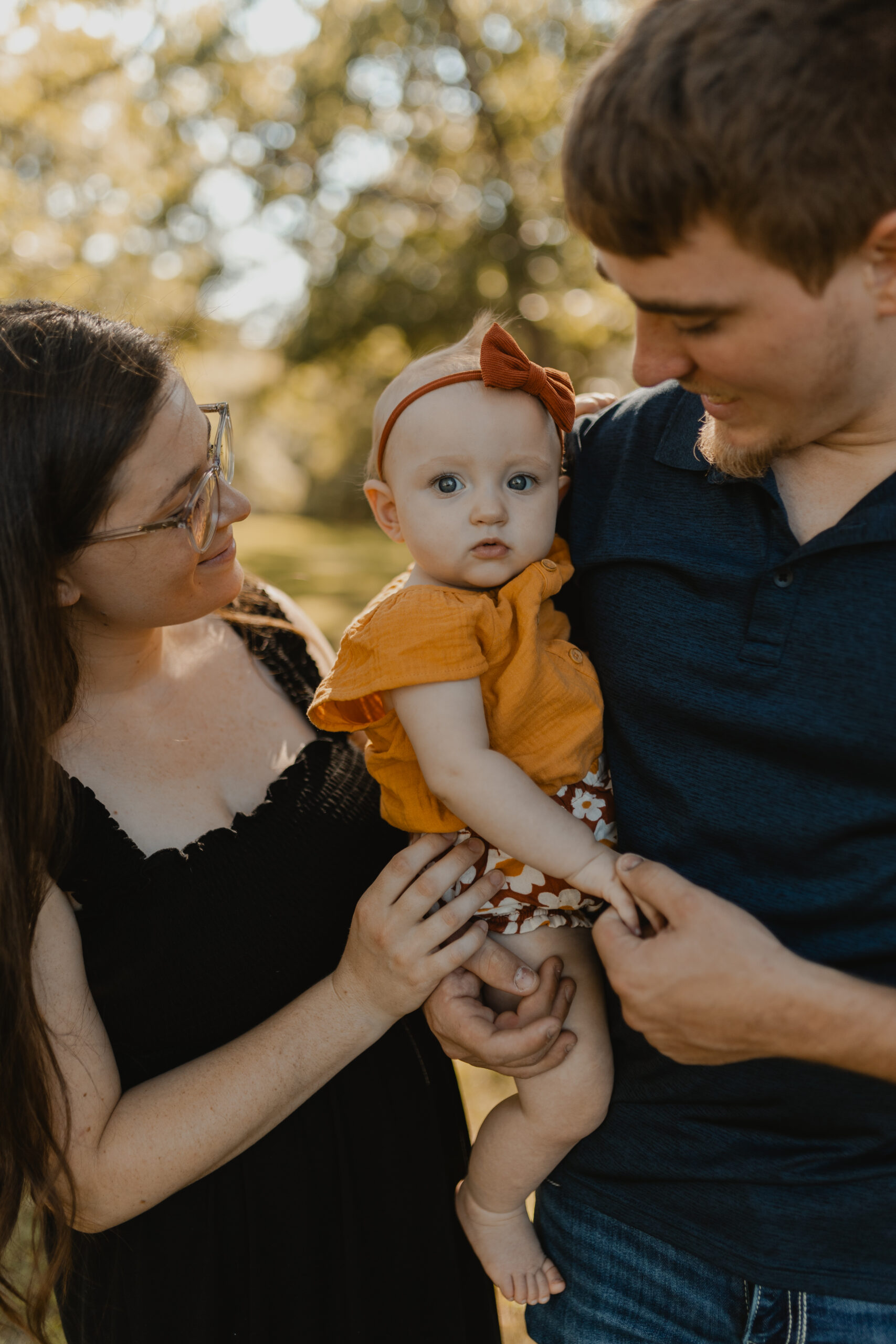 wife wearing black dress standing next to husband wearing navy shirt, holding baby girl in bright orange outfit 