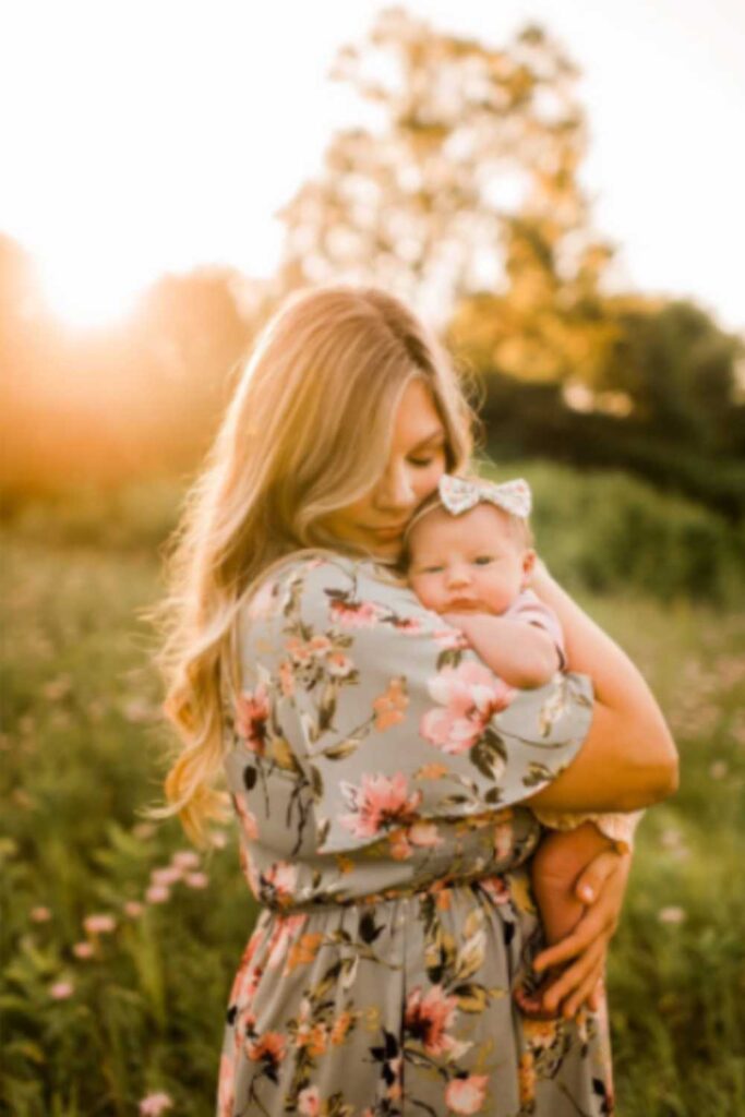 mom wearing floral green dress standing in field holding baby wearing pink outfit and bow