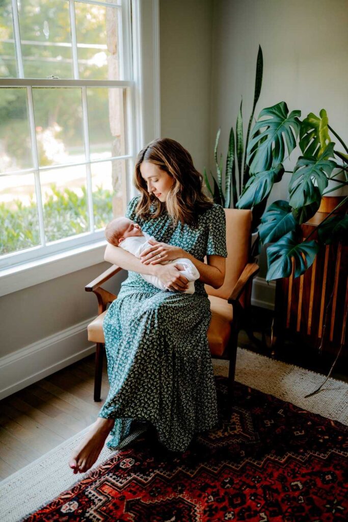 mom wearing long green dress sitting in rocking chair holding newborn baby in white blanket