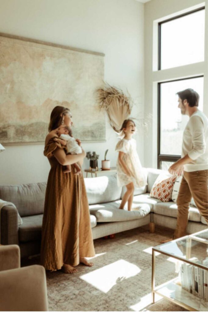 mom wearing burnt orange dress holding newborn in white blanket with a dad standing nearby interacting with young daughter jumping on couch