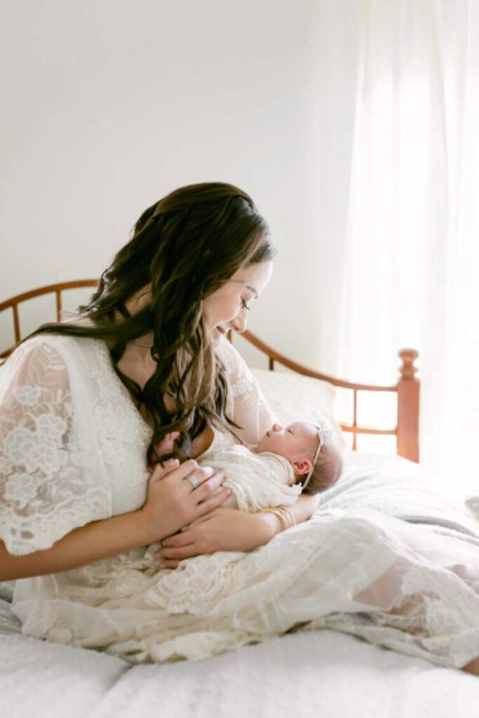 woman wearing white dress sitting on bed holding newborn baby in a white swaddle