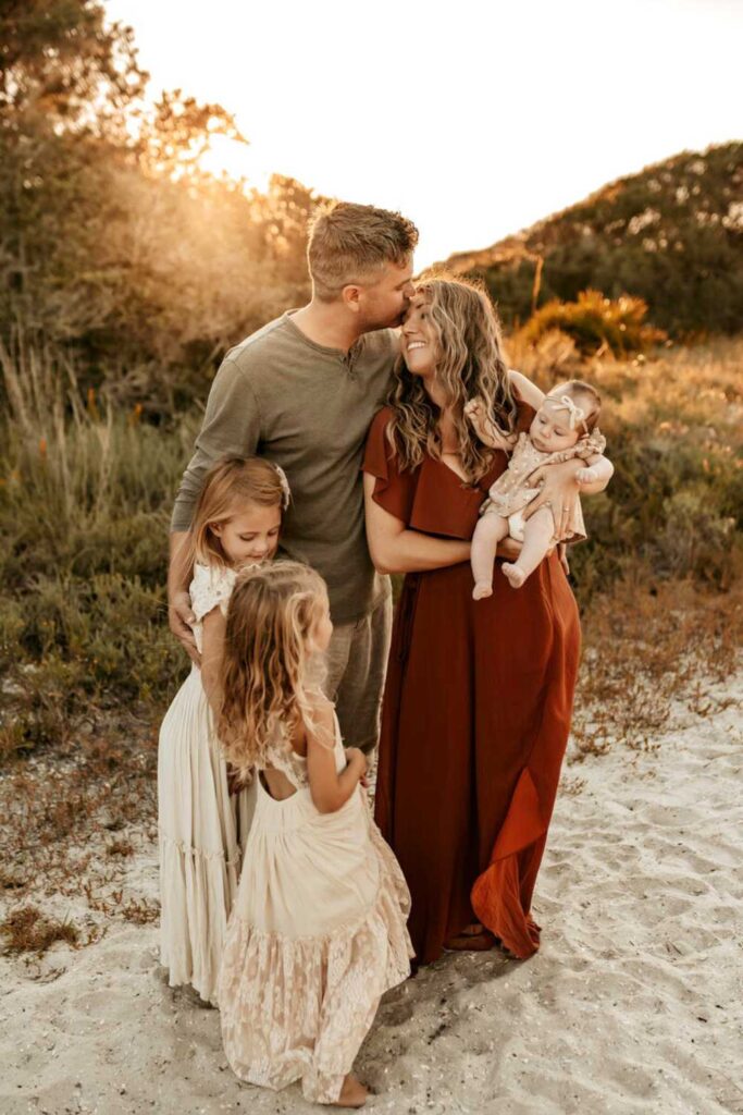 mom wearing red dress holding newborn and standing next to dad wearing green shirt with two daughters standing next to them in white dresses