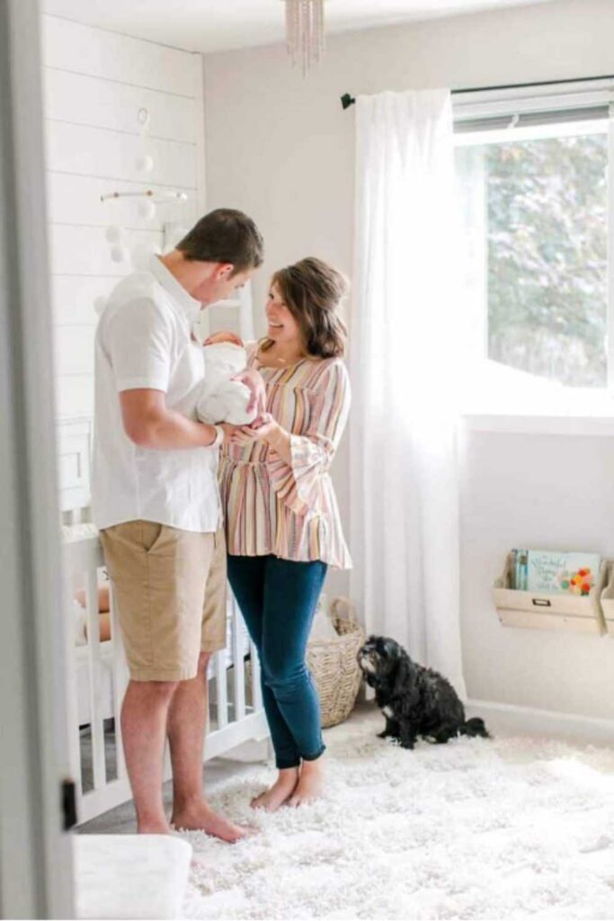 mom wearing striped shirt standing next to dad wearing white shirt holding baby wrapped in white blanket