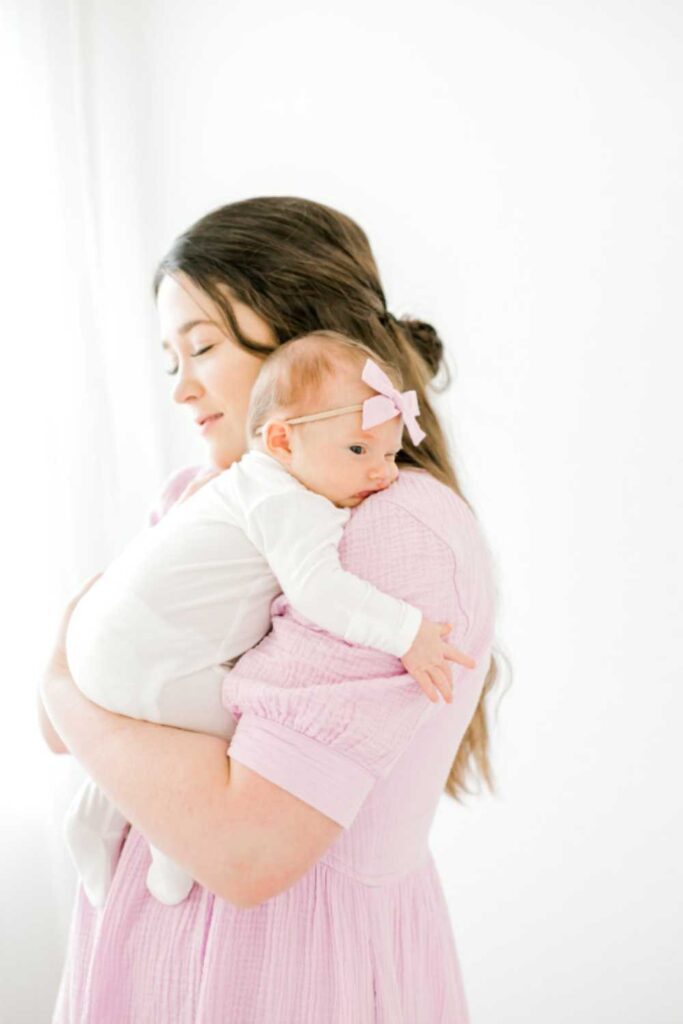 mom wearing pink dress holding newborn baby in white outfit